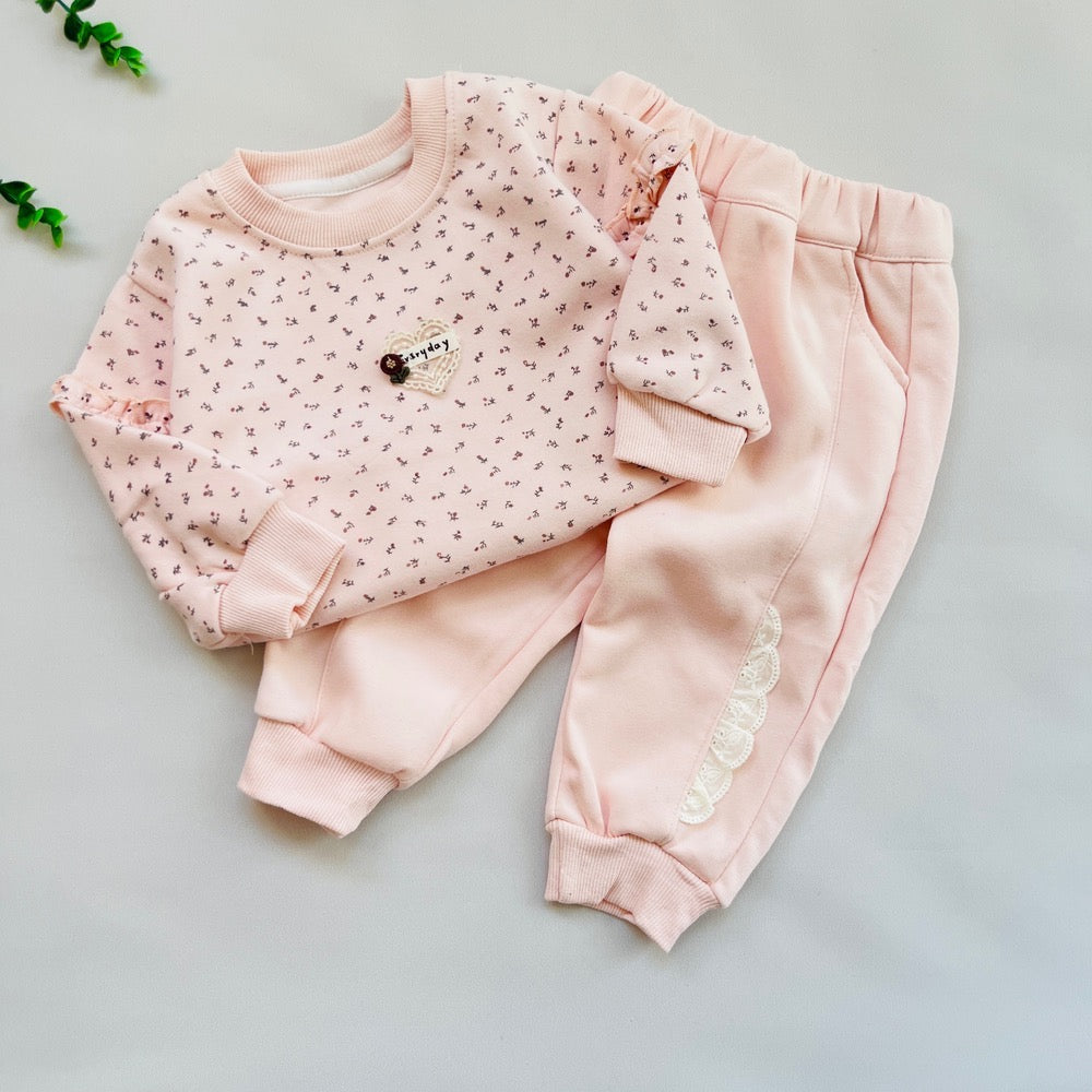 A two-piece baby clothing set laid flat on a light gray background, featuring a long-sleeve top and matching pants in pale pink. The top has a small allover delicate print and a small decorative chest emblem, while the pants have elastic cuffs and a decorative white scalloped detail near the lower leg.